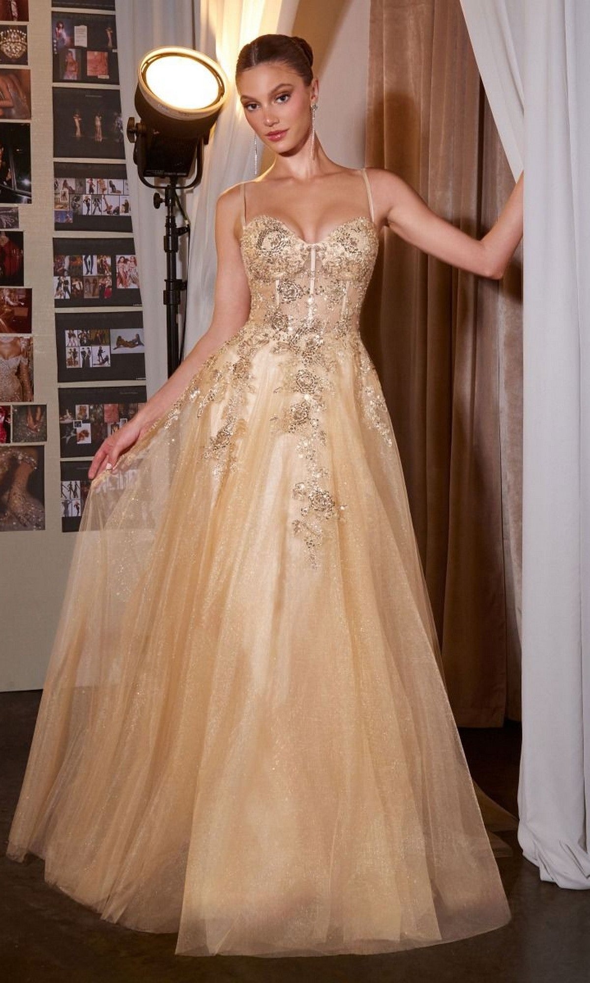A woman in the Ladivine C177, a gold floor-length prom dress with an embellished bodice, stands holding a curtain open in front of a light, with photo collages visible in the background.